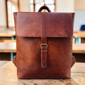 May include: A structured brown leather backpack featuring a fold-over flap closure secured by a vertical strap with a brass-toned metal buckle. The bag has a smooth, matte finish with visible natural grain texture and a top handle for carrying.