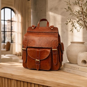 May include: A cognac-coloured leather backpack featuring a large fold-over flap, a front pocket with a buckle closure, two side pockets, and a top handle. The bag is accented with brass-toned metal hardware and visible stitching along the edges.