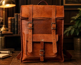 Può includere: Uno zaino in pelle color cuoio con patta superiore, due cinghie verticali con fibbie in metallo color ottone e una tasca frontale fissata da una cinghia centrale con fibbia. La borsa ha una forma rettangolare strutturata con cuciture.