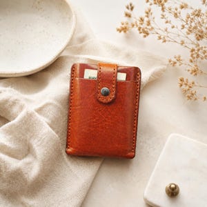 May include: A rectangular cognac-colored leather card holder featuring a vertical strap closure with a single metal snap button. The wallet has visible stitching along the edges and a top slot containing a small white card with green text.