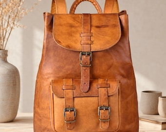 Può includere: Uno zaino in pelle color cuoio con un ampio scomparto principale dotato di patta, una cinghia verticale con fibbia in metallo e una tasca frontale fissata da due cinghie più piccole con fibbie in metallo, completo di manico e spallacci.