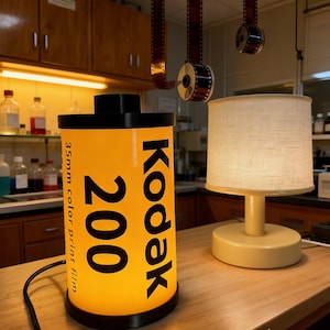 May include: A cylindrical lamp designed to resemble a Kodak 200 35mm color print film canister. The lamp features a bright yellow body with black text reading Kodak 200 35mm color print film, topped and bottomed with black circular caps.