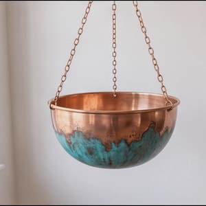 May include: A hanging bowl made of polished copper with a distinct turquoise patina finish on the lower exterior. The bowl is suspended by three thin copper chains that meet at a central point above the rim of the rounded vessel.