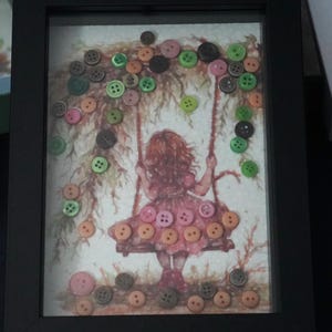 May include: A framed artwork featuring a girl on a swing with colorful buttons arranged around her. The buttons are in shades of green, pink, and brown, creating a whimsical and colorful scene.