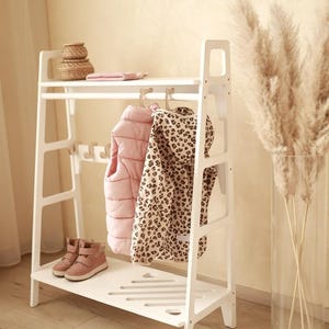 Peut inclure: Un portant à vêtements en bois blanc avec une étagère supérieure, une tringle et une étagère inférieure. Le portant supporte un gilet matelassé rose, une veste à capuche à motif léopard et une paire de baskets roses. Un petit panier tressé est posé sur l'étagère supérieure.