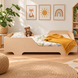 Op de afbeelding: Een lichtgekleurd houten peuterbed met lage zijhekken, voorzien van een wit laken met bloemenpatroon, een bruine teddybeer met een geruite strik en een textuurrijke mosterdgele deken gedrapeerd over het voeteneinde.