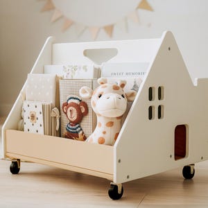 Könnte beinhalten: Ein weißes, hausförmiges Bücherregal auf Rollen mit einer Frontplatte aus hellem Holz. Das Regal enthält mehrere Bücher und ein Giraffen-Stofftier. Die Seite des Bücherregals weist ein ausgeschnittenes Fenster und ein kleines Bogentür-Design auf.