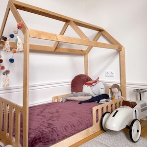 Op de afbeelding: Een lichtkleurig houten bedframe in de vorm van een huis met een pruimkleurige deken, diverse knuffels en een pompon-slinger. Een crèmekleurige speelgoedraceauto in vintage-stijl met het nummer één op de zijkant staat naast het bedframe.