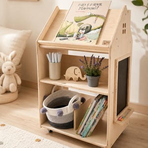 Könnte beinhalten: Ein helles, hausförmiges Bücherregal aus Holz auf Rollen mit einer oberen Ablage, zwei Innenregalen und einer seitlichen Tafel. Die Regale enthalten ein Buch mit dem Titel O QUE É O AMOR, MINIMONI?, einen Becher mit Stiften, einen Holzelefanten, eine Pflanze und einen Korb.