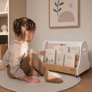 Könnte beinhalten: Ein weißes Bodenregal aus Holz mit Rollen, das über einen Boden in Naturholzoptik und seitliche Aussparungen verfügt. Das Regal enthält mehrere Bücher mit hellen Einbänden, darunter Titel wie My Little Adventures und Little Things.
