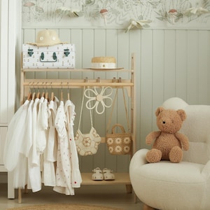 Peut inclure: Un portant à vêtements en bois clair supporte des vêtements blancs sur cintres, deux petits sacs en crochet et un accessoire en forme de fleur blanche. Un chapeau de paille, une malle de rangement à motifs et des chaussures blanches sont exposés.