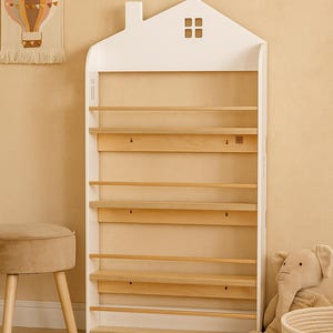 Könnte beinhalten: Ein weißes, hausförmiges Bücherregal aus Holz mit einem Schornstein und einem kleinen Fensterausschnitt an der Oberseite. Das Regal verfügt über vier horizontale Ablagen mit hellen, naturbelassenen Holzoberflächen und schmalen Frontleisten.