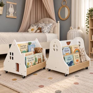 Könnte beinhalten: Zwei weiße, hausförmige Bodenbücherregale auf Rollen, jeweils mit ausgeschnittenen Fenstern und einem hellen Holzsockel. Die Regale sind mit Kinderbüchern mit den Titeln Good Night Little Bear, Little Explorer, The Very Brave Fox, Animal Friends, My First Poems, Colors, Little Ocean und Forest Friends sowie zwei Plüschbären gefüllt.
