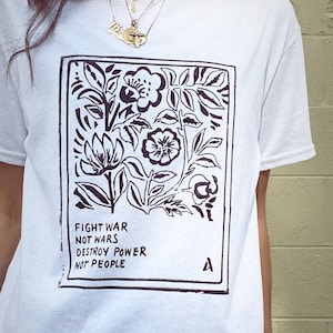 Peut inclure: T-shirt blanc avec un motif floral noir et le texte "FIGHT WAR NOT WARS DESTROY POWER NOT PEOPLE". Le motif est encadré en noir. Une petite lettre "A" est dans le coin inférieur droit.