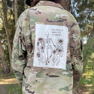 May include: A camouflage jacket with a white patch on the back that says "Fell asleep amid the flowers" and features a drawing of a skeleton in a field of sunflowers.