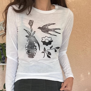 May include: White long-sleeved shirt with a black graphic print of a bird, flower, and a vase. The shirt is paired with a dark green pleated skirt.