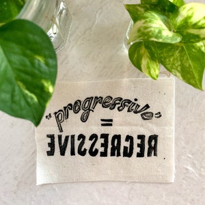 May include: A white fabric patch with black text that reads "progressive" in a curved font, followed by an equals sign and "regressive" in a mirrored font.