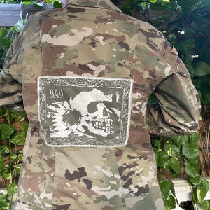 May include: A camouflage jacket with a white patch sewn on the back. The patch features a skull and crossbones design with the word "BAD" above it.