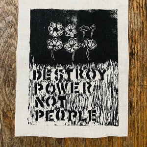 May include: A black and white fabric patch featuring a floral design above the text "DESTROY POWER NOT PEOPLE." The patch has a rectangular shape with a white border and is set against a wooden background.