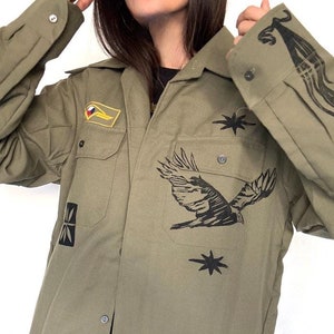 May include: A green utility shirt with black embroidered designs, including a bird, stars, and a patch with a flag. The shirt has a button-up front and long sleeves.