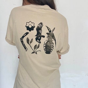 May include: A beige t-shirt with a black graphic print of a flower, a bird, a knife, leaves, and a grenade.