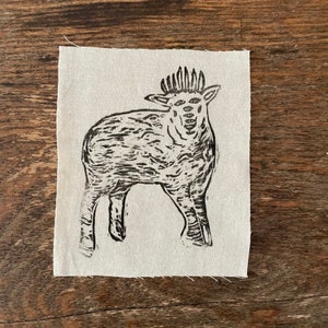 May include: A black and white linocut print of a stylised sheep with multiple eyes on a beige fabric background.