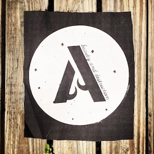May include: Black fabric patch with a white circle containing a stylised letter A and the text "beauty and destruction".