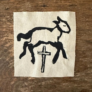 May include: Black and white illustration of a lamb standing on a cross. The image is printed on a beige fabric.