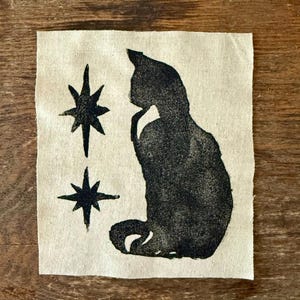 Puede incluir: Silueta negra de un gato y dos estrellas impresas en un cuadrado de tela beige. El gato está sentado, con la barbilla apoyada en la pata. Las estrellas están situadas a la izquierda del gato.