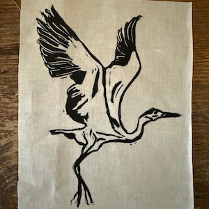 Puede incluir: Un grabado en linóleo en blanco y negro de un pájaro en vuelo, con las alas extendidas y un cuello largo. El pájaro está representado de forma estilizada y artística. La impresión está sobre un trozo de tela de color claro, probablemente lino o algodón.