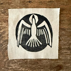 May include: A black and white print of a dove with outstretched wings, centered in a circle on a beige fabric background.