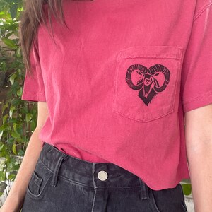 May include: A short-sleeved, dusty rose t-shirt with a pocket featuring a black ram graphic. The shirt is paired with black denim shorts. The ram graphic is centered on the pocket.