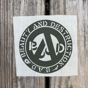 May include: A white fabric patch with a dark green circle containing a stylized anarchy symbol and the text "Beauty and Destruction."
