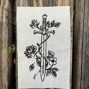 Peut inclure: Un écusson rectangulaire avec une illustration noire d'une épée entrelacée de roses et de feuilles. Le dessin est imprimé sur un tissu beige clair. Les initiales "BAD" sont imprimées sur l'épée.