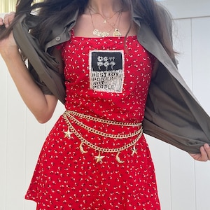 May include: Red floral print romper with a black and white patch that says "Destroy Power Not People". The romper has a gold chain belt with star and crescent moon charms.