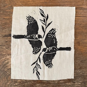 May include: A square, off-white fabric patch featuring a black linocut print of two birds in flight, facing each other. The birds are positioned around a sprig of leaves. The print has a rustic, handmade aesthetic.