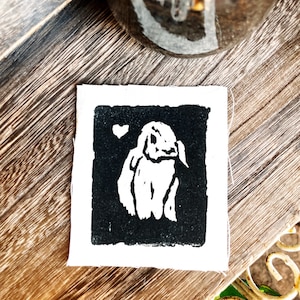 May include: A black and white linocut print of a rabbit on a white background. The rabbit is in a sitting position with its ears up and its eyes closed. The print is framed by a white border.