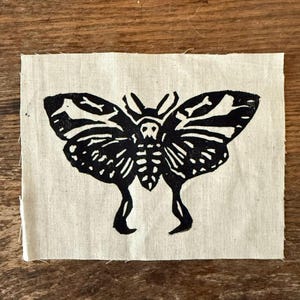 May include: Black and white linocut print of a moth with a skull design on a square piece of natural-colored fabric. The moth has detailed wing patterns and a skeletal design. The fabric has a raw, unfinished edge.
