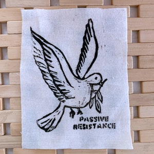 Könnte beinhalten: Ein weißes Stoffpatch mit einem schwarzen Linolschnitt einer Taube, die einen Olivenzweig trägt. Die Worte "PASSIVE RESISTANCE" sind darunter in Schwarz gedruckt. Der Patch hat einen rohen Rand.