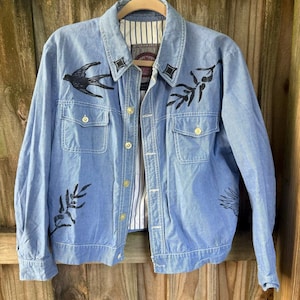 May include: Light blue denim jacket with black bird and floral designs. The jacket has two chest pockets, gold buttons, and a striped lining visible at the collar and cuffs. The jacket is hanging on a wooden hanger.