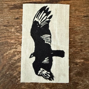 May include: A black and white linocut print of a bird in flight, possibly an eagle or hawk, is centered on a rectangular piece of natural-colored fabric. The bird's wings are outstretched, and the linocut style gives it a textured appearance.