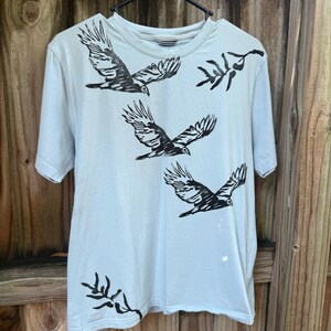 May include: A light blue short-sleeved t-shirt featuring a black ink-style print of three birds in flight and two leafy branch motifs. The garment has a crew neckline and is displayed on a black hanger against a wooden background.