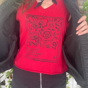 Peut inclure: T-shirt rouge à col en V avec un motif floral noir et le texte "Fight war not wars destroy power not people".