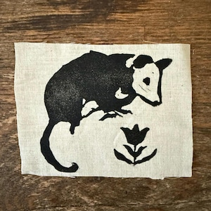 May include: A black and white linocut print of an opossum and a flower on a rectangular piece of natural-colored fabric. The opossum is depicted in profile, with its tail curled. The flower is a simple, stylized design.