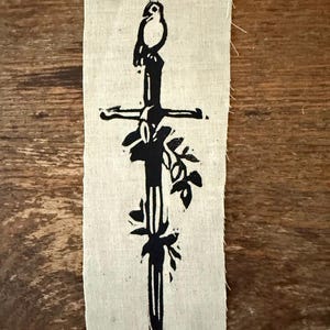 Op de afbeelding: Een zwart-wit linoleumsnede van een zwaard met een vogel erbovenop en bloemaccenten. De print staat op een rechthoekig stuk stof in natuurlijke kleur met gerafelde randen. Het kunstwerk wordt op een houten oppervlak gepresenteerd.
