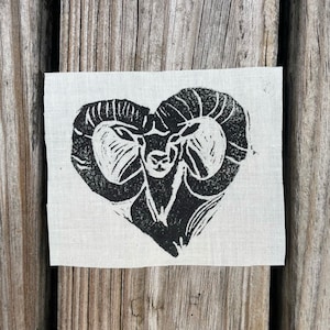 May include: Black and white print of a ram's head with large horns on a white linen fabric background.