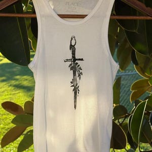 May include: White ribbed tank top with a black sword design and floral accents. The tank top is hanging on a wooden hanger. The brand name "PLACE" is visible on the neckline.