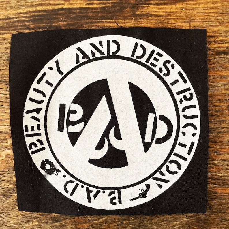 Puede incluir: Parche de tela negra con un dise&ntilde;o circular blanco. El dise&ntilde;o presenta las palabras "BEAUTY AND DESTRUCTION" alrededor del per&iacute;metro, con "B.A.D." en la parte inferior. Una "A" estilizada y otros s&iacute;mbolos est&aacute;n en el centro.