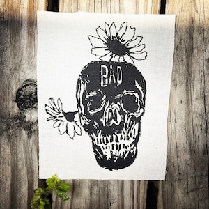 May include: Black and white illustration of a skull with the word "BAD" printed on its forehead. The skull is wearing a flower crown.