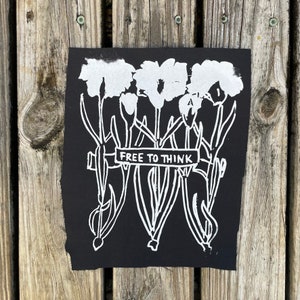 May include: Black fabric patch with a white design of three stylized flowers with stems and leaves. The flowers are tied together with a banner that reads "FREE TO THINK".